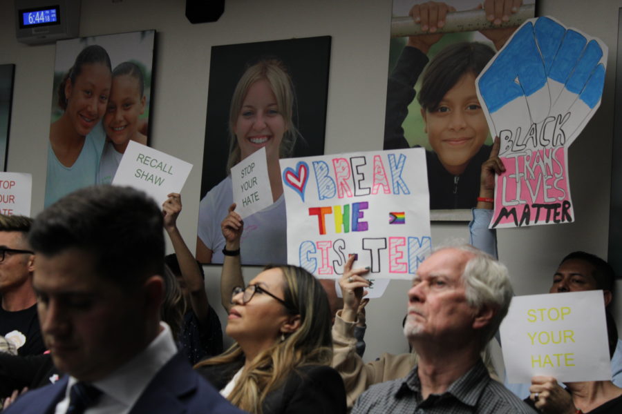 Assemblymember Bill Essayli (foreground, left) sits in the audience as protestors in attendance hold up signs.
