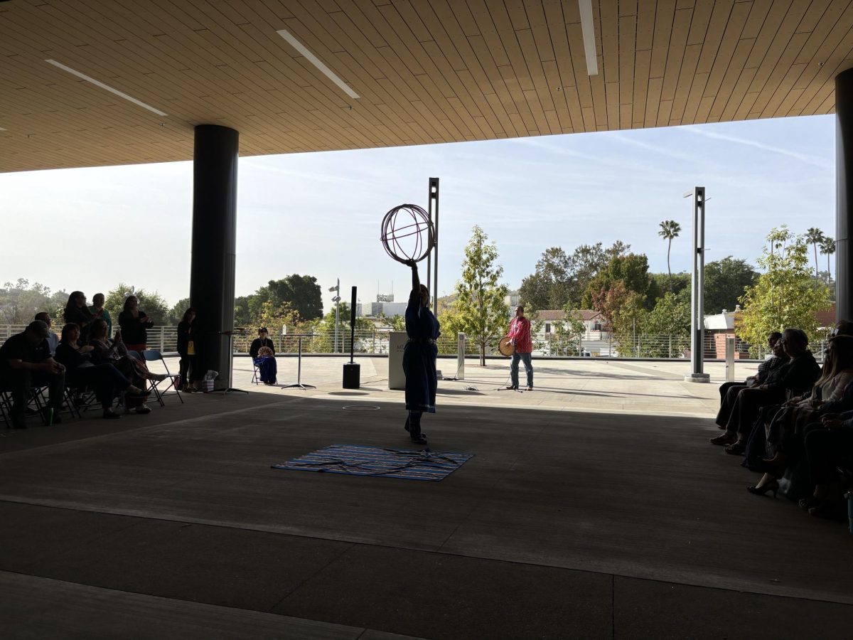 Tahnaban Whipple in a traditional Navajo (Diné) dress and her father concluding the hoop dance. To the left is Tahnaban’s mother holding a traditional basket used by the Diné people in ceremonies.